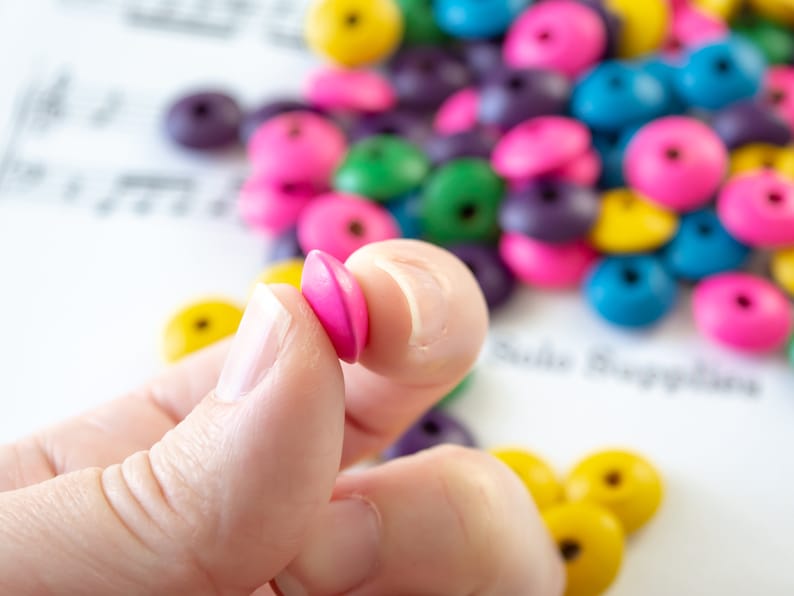 May include: A hand holding two pink wooden beads. The beads are round and have a smooth surface. The beads are in focus, while the background is blurred. The background includes a variety of colorful wooden beads.