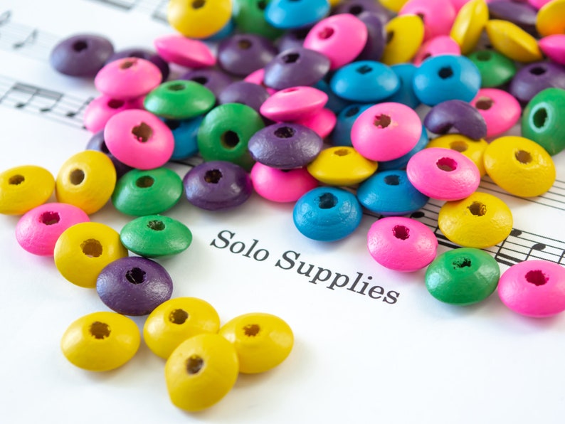 May include: A collection of colorful wooden beads in various shades of pink, blue, green, yellow, and purple. The beads are scattered on a white surface with the text "Solo Supplies" printed on it.