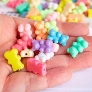 May include: A hand holding a variety of colorful gummy bear-shaped beads. The beads are made of plastic and are in shades of pink, yellow, blue, green, and purple.