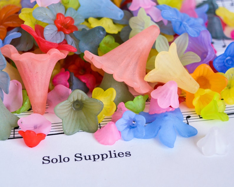 May include: Assortment of colourful plastic flower-shaped beads in various shades of pink, blue, yellow, green, and orange. The beads are scattered on a white surface with musical notes printed on it. The text "Solo Supplies" is printed on the surface.