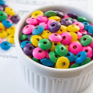 May include: A white ceramic bowl filled with colorful wooden beads. The beads are in shades of pink, blue, green, yellow, and purple.