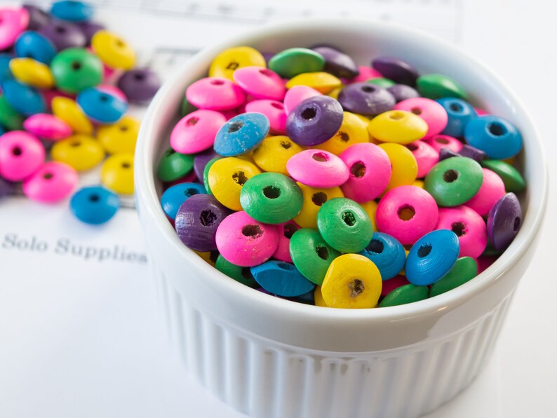 May include: A white ceramic bowl filled with colorful wooden beads. The beads are in shades of pink, blue, green, yellow, and purple.