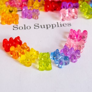May include: A rainbow assortment of gummy bear charms, arranged in a semi-circle on a white surface. The gummy bears are made of clear plastic and are in various colors, including red, orange, yellow, green, blue, purple, and pink. The text "Solo Supplies" is printed in black on the surface.