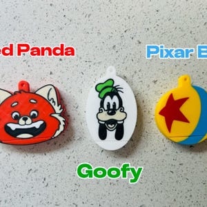 May include: Three colorful keychains featuring cartoon characters. The first keychain is red and features a red panda face. The second keychain is white and features Goofy. The third keychain is yellow and blue and features a Pixar ball with a red star.