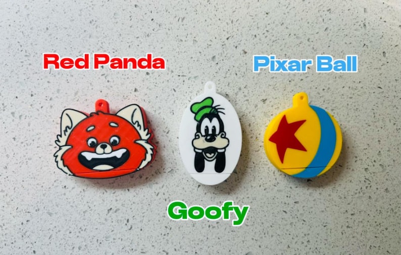 May include: Three colorful keychains featuring cartoon characters. The first keychain is red and features a red panda face. The second keychain is white and features Goofy. The third keychain is yellow and blue and features a Pixar ball with a red star.