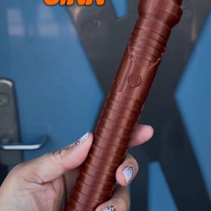 K&ouml;nnte beinhalten: Ein brauner Lichtschwertgriff mit dem Text "QUI-GON JINN" in orangefarbenen Buchstaben.