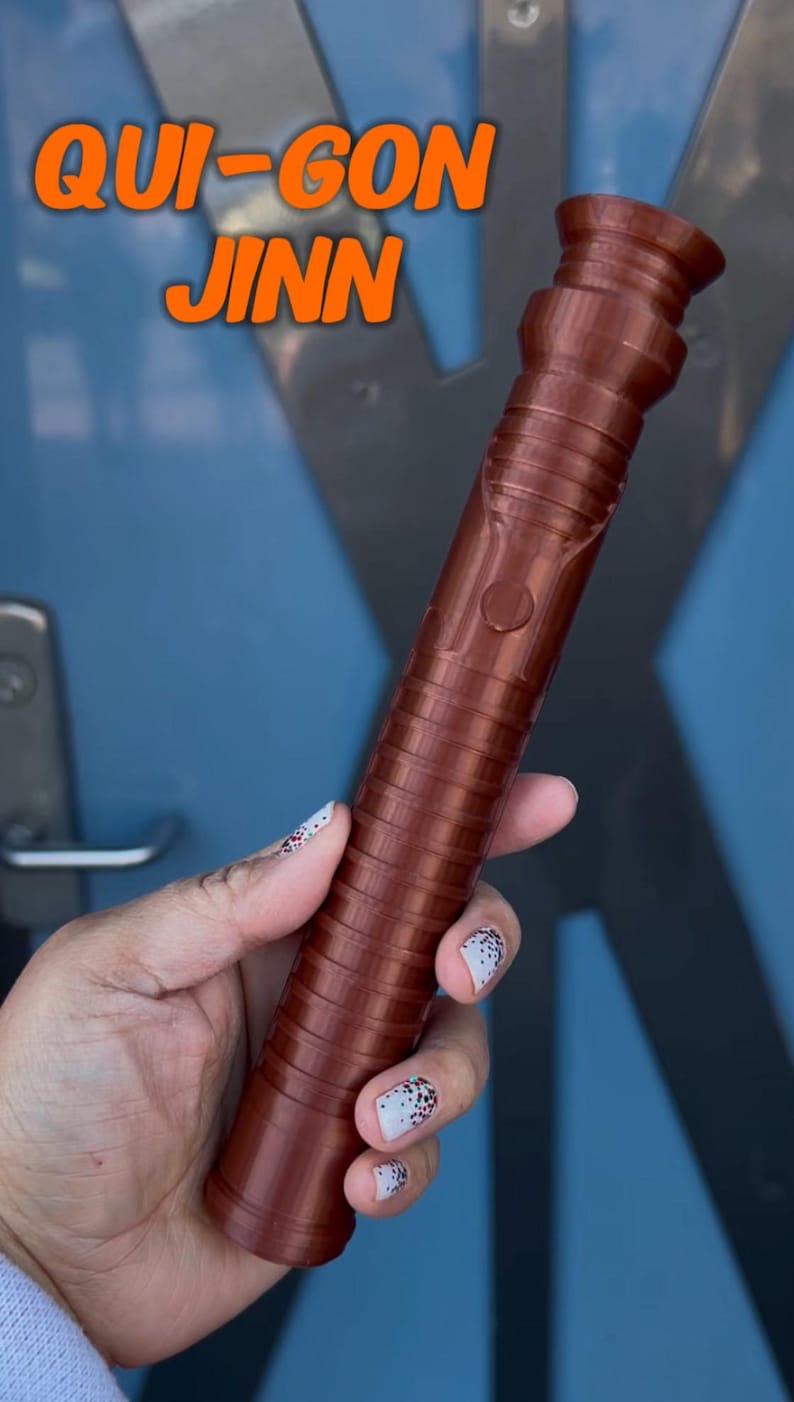 K&ouml;nnte beinhalten: Ein brauner Lichtschwertgriff mit dem Text "QUI-GON JINN" in orangefarbenen Buchstaben.
