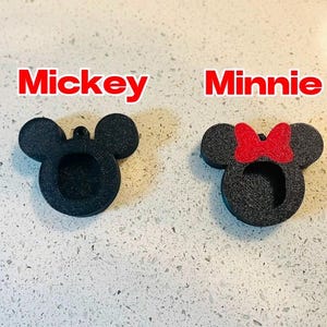 May include: Two black foam cutouts of Mickey Mouse and Minnie Mouse. Mickey Mouse has large round ears and Minnie Mouse has a red bow on her head.