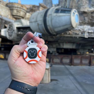 May include: A white and orange BB-8 keychain with a black Mickey Mouse silhouette on the front. The keychain is being held in a person's hand in front of a Star Wars themed attraction.