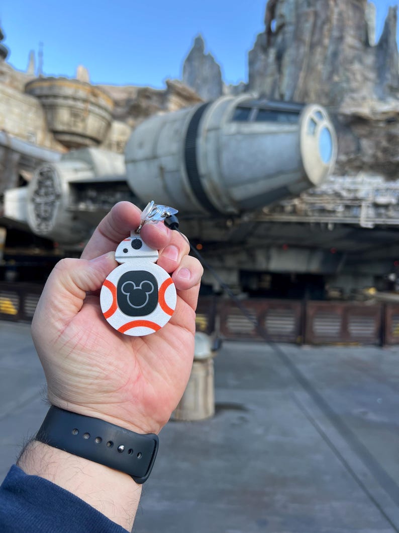 May include: A white and orange BB-8 keychain with a black Mickey Mouse silhouette on the front. The keychain is being held in a person's hand in front of a Star Wars themed attraction.