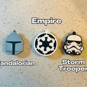 May include: Three white and black keychains featuring Star Wars characters. The keychains are shaped like the helmets of a Mandalorian, a Stormtrooper, and the Empire symbol.