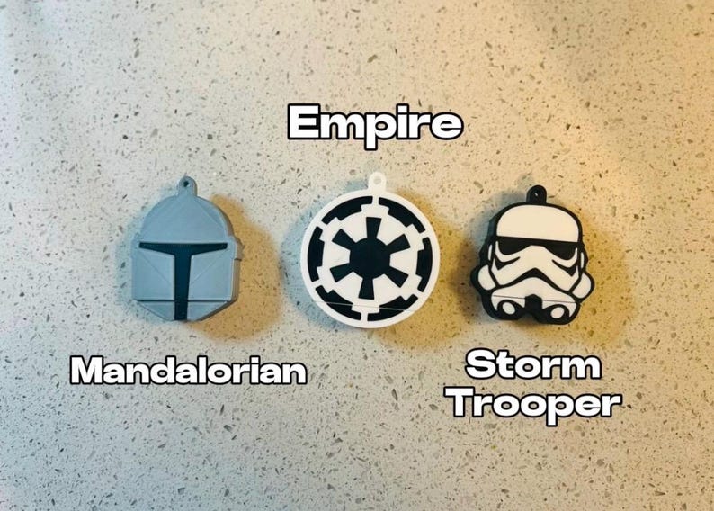 May include: Three white and black keychains featuring Star Wars characters. The keychains are shaped like the helmets of a Mandalorian, a Stormtrooper, and the Empire symbol.