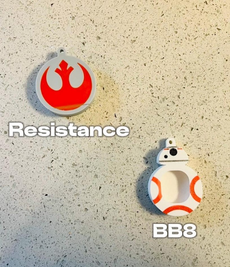 May include: Two round, white, 3D printed ornaments. The first ornament features the red Rebel Alliance symbol with the text "Resistance". The second ornament features a white and orange BB-8 character with the text "BB8".