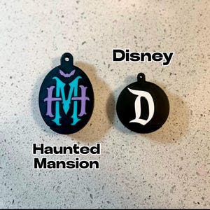 May include: Two black round ornaments with white lettering. The first ornament has the text "Haunted Mansion" and a purple and teal monogram. The second ornament has the text "Disney" and a white "D".
