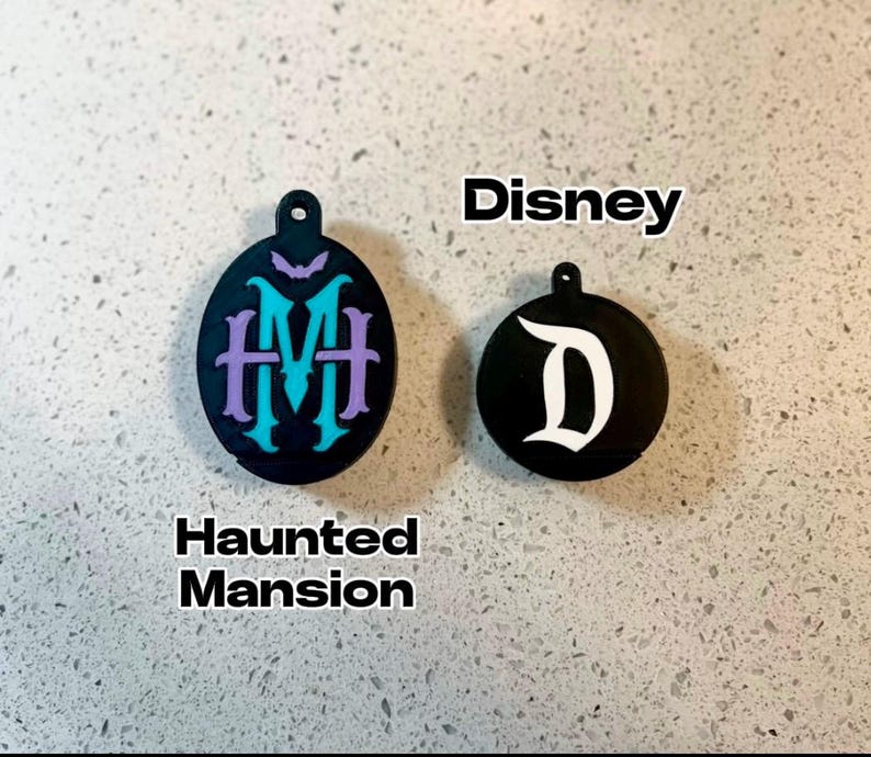 May include: Two black round ornaments with white lettering. The first ornament has the text "Haunted Mansion" and a purple and teal monogram. The second ornament has the text "Disney" and a white "D".
