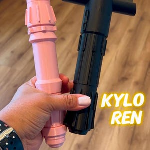 K&ouml;nnte beinhalten: Zwei Lichtschwert-Requisiten, eine rosa und eine schwarze, mit den Worten "REY" und "KYLO REN" darauf.