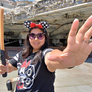 K&ouml;nnte beinhalten: Eine Frau tr&auml;gt ein schwarz-wei&szlig;es Shirt mit einem roten Minnie Mouse-Motiv und ein rot-wei&szlig; gepunktetes Minnie Mouse-Haarband. Sie h&auml;lt einen braunen Churro vor einem gro&szlig;en Raumschiff.