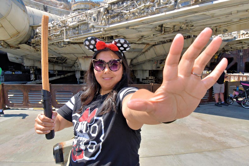 K&ouml;nnte beinhalten: Eine Frau tr&auml;gt ein schwarz-wei&szlig;es Shirt mit einem roten Minnie Mouse-Motiv und ein rot-wei&szlig; gepunktetes Minnie Mouse-Haarband. Sie h&auml;lt einen braunen Churro vor einem gro&szlig;en Raumschiff.