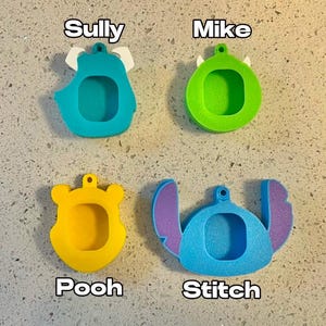 May include: Four different character-shaped keychains, each with a blank space for a photo. The keychains are blue, green, yellow, and light blue. The characters are Sully, Mike, Pooh, and Stitch.