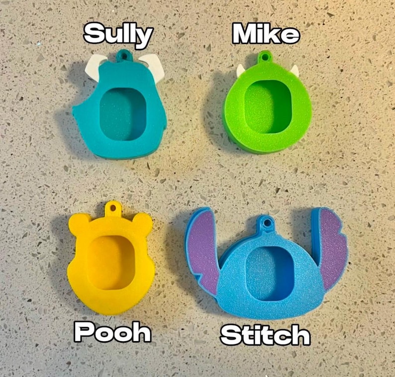 May include: Four different character-shaped keychains, each with a blank space for a photo. The keychains are blue, green, yellow, and light blue. The characters are Sully, Mike, Pooh, and Stitch.