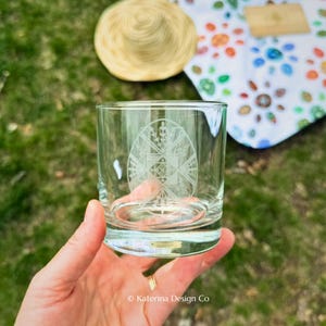Puede incluir: Un vaso de vidrio transparente con un diseño grabado de un huevo adornado. El vaso se sostiene en una mano, con un sombrero de paja y una manta colorida en el fondo. El texto "Katerina Design Co" está en la parte inferior.