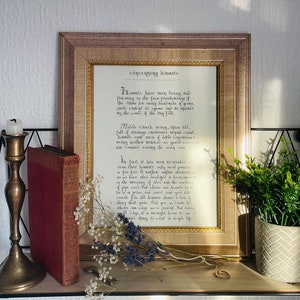 May include: A framed print with a gold border and a black and white handwritten text about hobbits. The text reads "Hobbits have been living and farming in the four farthings of the Shire for many hundreds of years, quite content to ignore and be ignored by the world of the big folk."