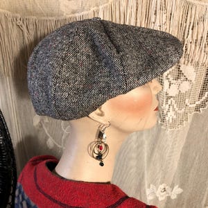May include: A gray tweed newsboy cap with a rounded crown and a short, stiff brim. The cap has a textured appearance with flecks of red and blue. The cap is worn by a mannequin with a red and blue patterned top.