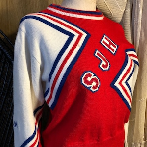 May include: A red and white long-sleeve knit sweater featuring blue, white, and red striped chevron accents across the chest and sleeves. The letters SJH are embroidered in red with a white outline on the front. The collar has a ribbed trim.
