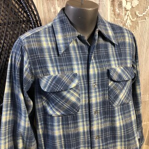 May include: A long-sleeve button-down shirt featuring a plaid pattern in shades of blue, grey, and pale yellow. The shirt includes a pointed collar, a full-length front button closure, and two chest pockets with buttoned flaps on each side.