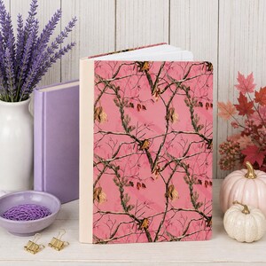 May include: A hardcover notebook featuring a vibrant pink background with a repeating camouflage pattern of brown tree branches and green pine needles. The spine of the notebook is a solid cream color, and the pages are visible at the top edge.