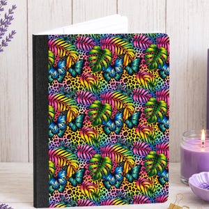 May include: A notebook featuring a vibrant, multicolored pattern of tropical monstera leaves, palm fronds, and butterflies set against a yellow and black leopard print background. The notebook has a solid black spine and white interior pages.
