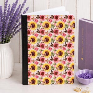 May include: A notebook with a black spine and a cover featuring a repeating pattern of yellow sunflowers, pink flowers, and small brown butterflies. The notebook is standing upright, showing its front cover design with a white interior page edge.