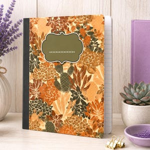 May include: A notebook with a black spine and a cover featuring a repeating pattern of orange, brown, and dark green succulents and cacti. A solid olive green label with a dotted line for writing is centered on the front cover of the notebook.