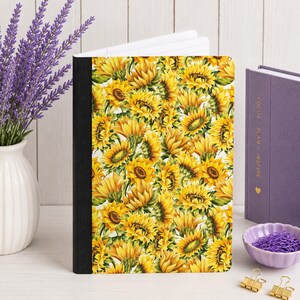 May include: A hardcover journal featuring a vibrant, repeating pattern of yellow sunflowers with green leaves on a white background, accented by a solid black spine. The cover displays a dense, detailed illustration of numerous blooming sunflowers.