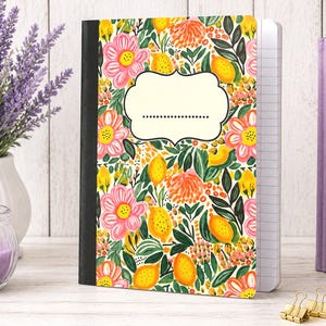 May include: A composition notebook with a black spine and a cover featuring a vibrant pattern of yellow lemons, pink flowers, and orange blossoms on a white background, including a central cream-colored label with a dotted line for writing.