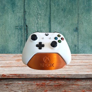 May include: A white Xbox controller is sitting on a copper-colored stand with the word "XBOX" printed on it. The stand is on a wooden surface.