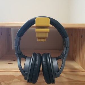 Op de afbeelding: Een zwarte koptelefoon met een gele plastic koptelefoonhouder bevestigd aan een houten plank.