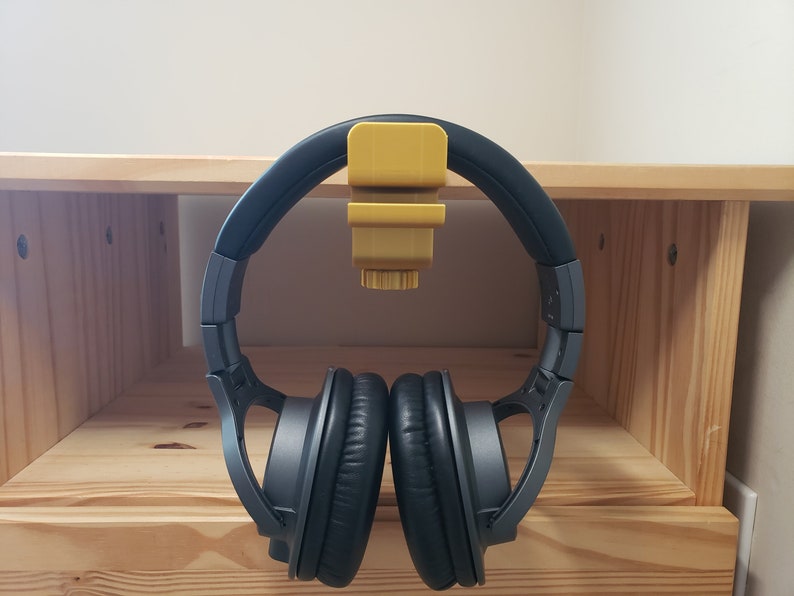 Op de afbeelding: Een zwarte koptelefoon met een gele plastic koptelefoonhouder bevestigd aan een houten plank.
