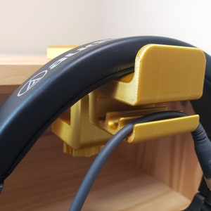 Op de afbeelding: Een gouden 3D-geprinte koptelefoonstandaard met een kabelhouder. De standaard houdt een zwarte koptelefoon met een zwarte kabel vast.