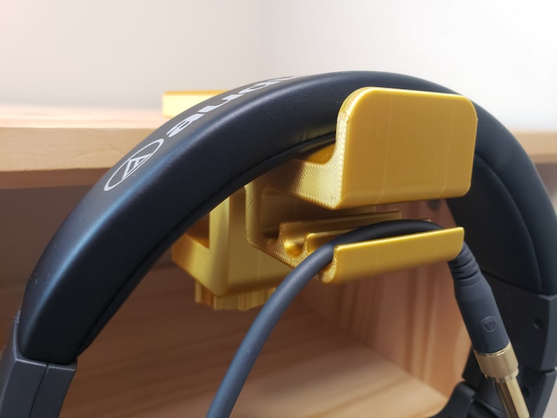 Op de afbeelding: Een gouden 3D-geprinte koptelefoonstandaard met een kabelhouder. De standaard houdt een zwarte koptelefoon met een zwarte kabel vast.