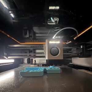 Może przedstawiać: Drukarka 3D w trakcie pracy, drukująca jasnoniebieski obiekt. Głowica drukująca jest beżowa i ma napis "Bambu Lab". Maszyna jest zamknięta w ciemnej obudowie. Obiekt jest w trakcie budowy.