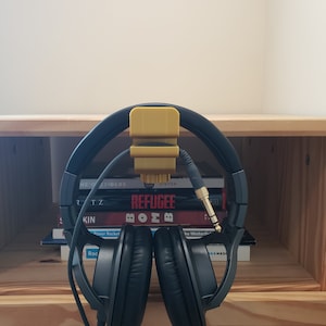 Op de afbeelding: Een zwarte over-ear koptelefoon met een gele koptelefoonhouder bevestigd aan een plank. De koptelefoon hangt aan de houder en de kabel is om de koptelefoon gewikkeld. De koptelefoon rust op een stapel boeken met titels als "The Outsiders", "Refugee" en "Bomb".