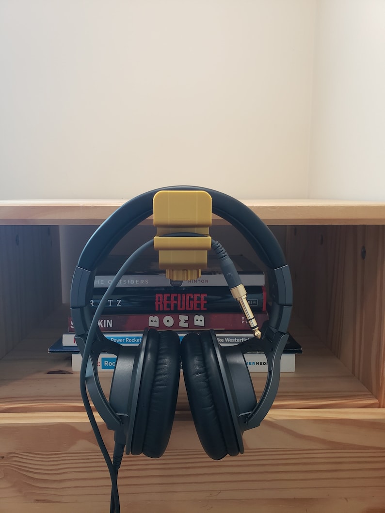 Op de afbeelding: Een zwarte over-ear koptelefoon met een gele koptelefoonhouder bevestigd aan een plank. De koptelefoon hangt aan de houder en de kabel is om de koptelefoon gewikkeld. De koptelefoon rust op een stapel boeken met titels als "The Outsiders", "Refugee" en "Bomb".
