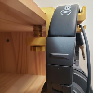 Op de afbeelding: Een gouden metalen hoofdtelefoonstandaard met een klem die aan een houten plank wordt bevestigd. De standaard houdt een zwarte hoofdtelefoon vast met de merknaam "Audio-Technica" gedrukt op de oorschelpen.