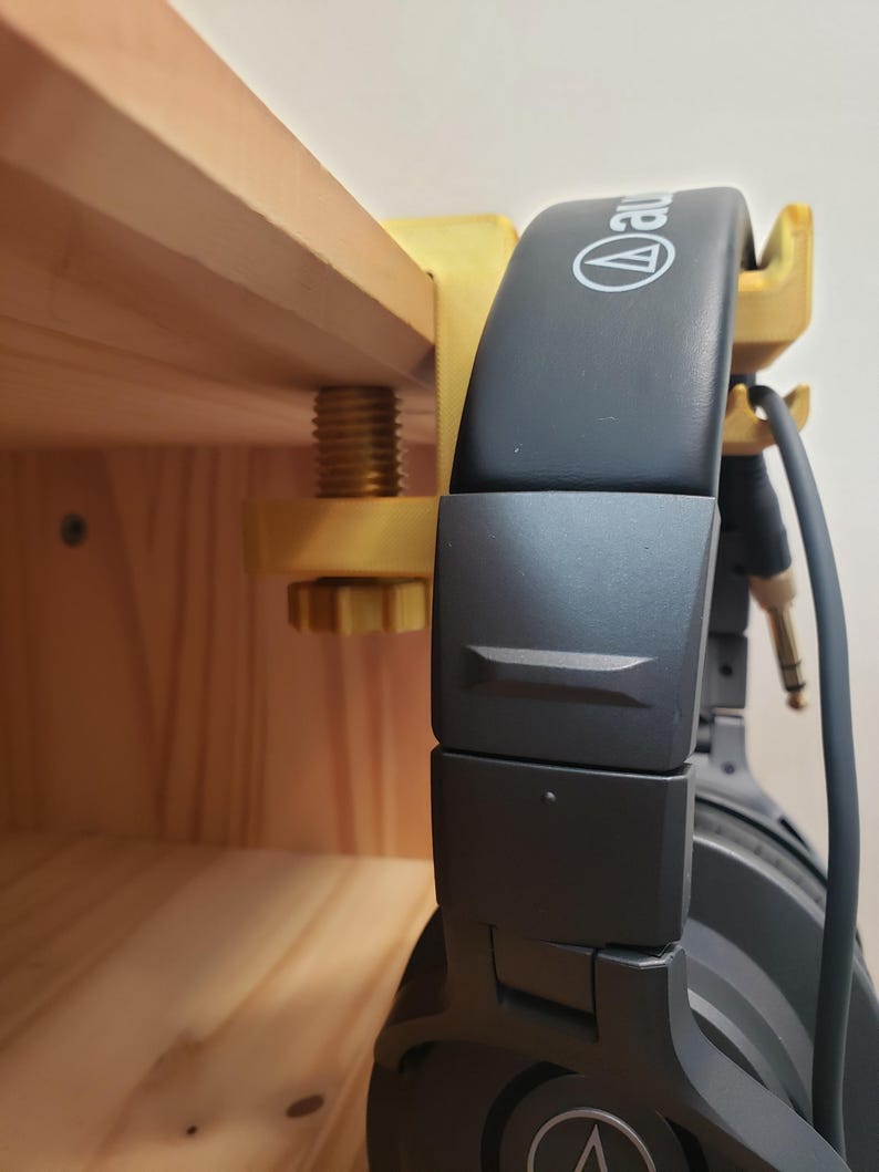 Op de afbeelding: Een gouden metalen hoofdtelefoonstandaard met een klem die aan een houten plank wordt bevestigd. De standaard houdt een zwarte hoofdtelefoon vast met de merknaam "Audio-Technica" gedrukt op de oorschelpen.