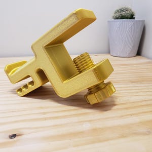 Op de afbeelding: Een gouden metalen klem met een schroefknop. De klem is ontworpen om voorwerpen veilig vast te houden.