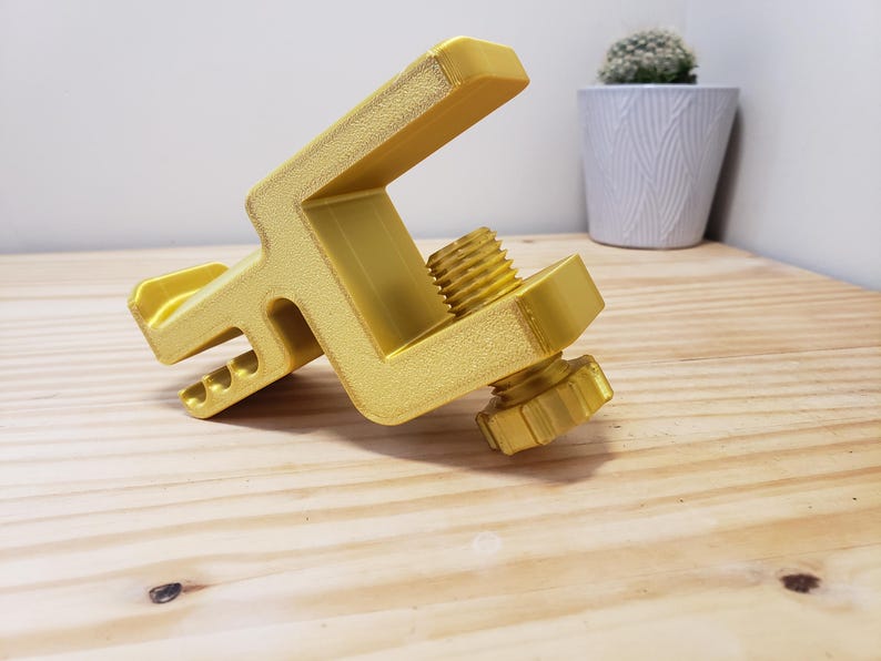Op de afbeelding: Een gouden metalen klem met een schroefknop. De klem is ontworpen om voorwerpen veilig vast te houden.