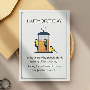 May include: A birthday card with the text "HAPPY BIRTHDAY" and a humorous message about aging and birds. The card features a bird feeder illustration with three birds, a tan envelope, and scissors.