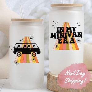 May include: Two white tumblers with lids and straws. One tumbler features a black silhouette of a minivan driving on a rainbow road with the text "In My Minivan Era". The other tumbler features a black silhouette of a minivan driving on a rainbow road with flowers.