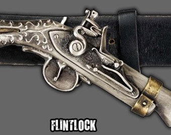 May include: A silver and gold flintlock pistol belt buckle with intricate engravings. The buckle is attached to a black leather belt.